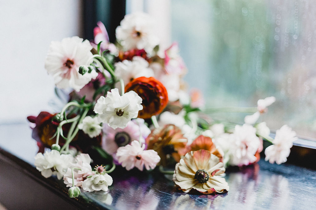 Soft white and warm-toned flowers scattered on a dark surface by a rain-speckled window